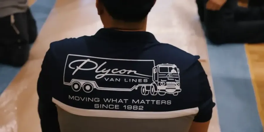 A person wearing a navy blue and grey Plycon Van Lines uniform shirt featuring a logo of a moving truck and the slogan "Moving What Matters Since 1982."